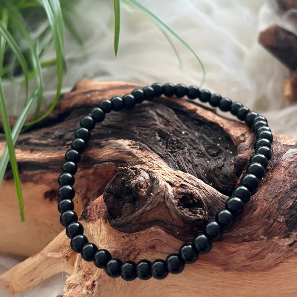 Ein elastisches Armband aus glänzend schwarzen Obsidian-Perlen liegt auf einem naturbelassenen Holzstück, umgeben von weichem, weißem Untergrund und frischem Grün – ein kraftvoller Schutzstein zur Erdung, zur Abwehr negativer Energien und zur Stärkung der inneren Klarheit.