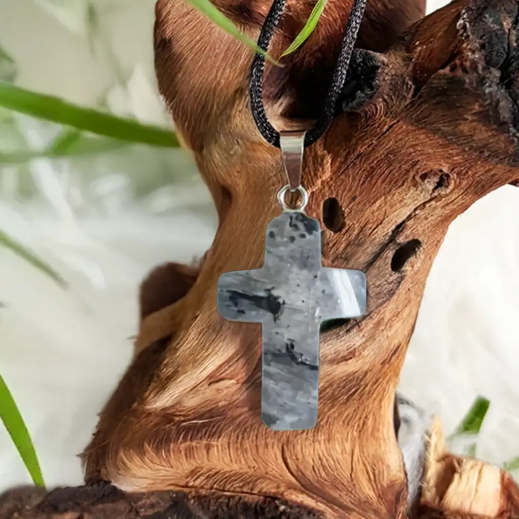 Detailaufnahme eines Labradorit Kreuz Anhängers mit faszinierendem Farbspiel, aufgehängt vor natürlichem Holz- und Pflanzenhintergrund