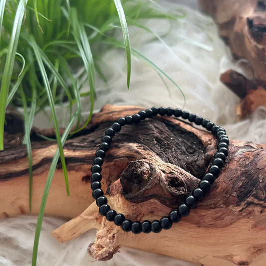Ein elastisches Armband aus glänzend schwarzen Obsidian-Perlen liegt auf einem naturbelassenen Holzstück, umgeben von weichem, weißem Untergrund und frischem Grün – ein kraftvoller Schutzstein zur Erdung, zur Abwehr negativer Energien und zur Stärkung der inneren Klarheit